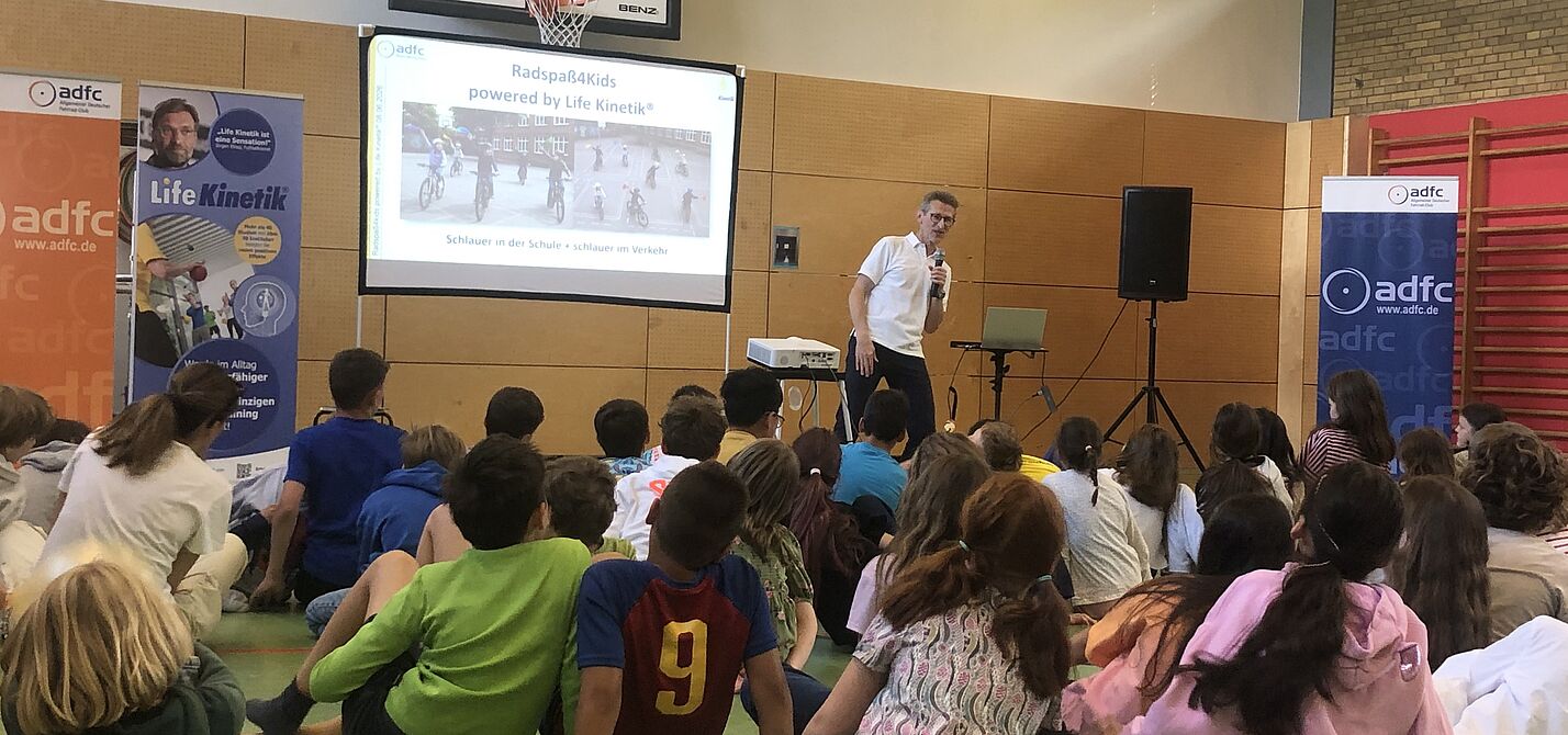 Life Kinetik®-Gründer Horst Lutz zeigt Bewegungsübung vor Klasse in Turnhalle vor. Er und die Kinder beugen sich nach rechts.