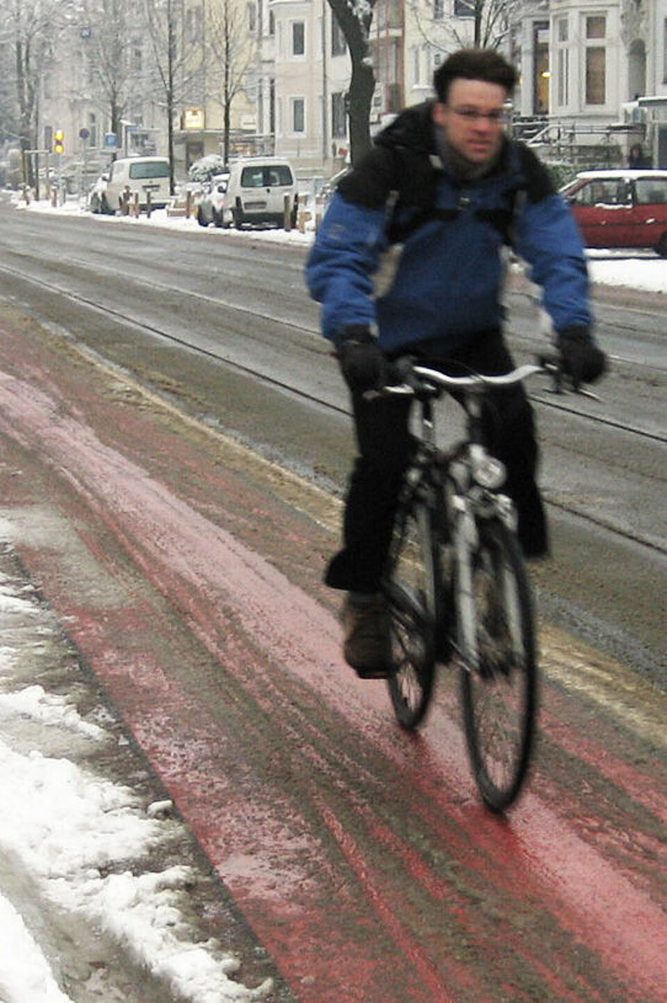 Radfahren im Winter Ein Radfahrer in Winterkleidung fährt auf einem rot markierten Radfahrstreifen neben einer Straße, der weitgehend von Schnee befreit ist, während am Straßenrand und auf geparkten Autos Schnee liegt.